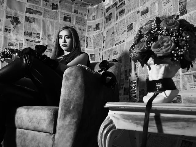VenusBarret with long hair, dark lipstick, black leather pants, and boots, sitting on a velvet chair, looking seductive, next to a bouquet and a mirror. Black and white.
