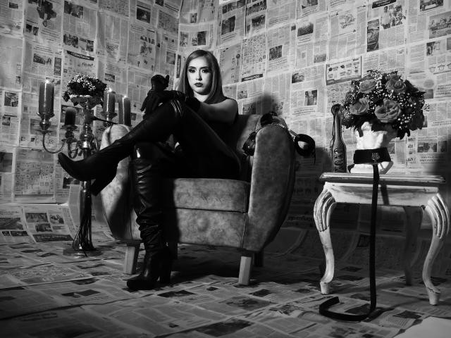 VenusBarret with her tits covered, sitting in a chair, wearing black thigh-high boots, legs crossed, in a newspaper-covered room, with a candelabrum and flower vase on a table.