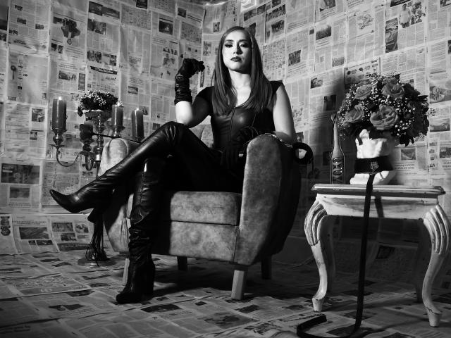 VenusBarret, with black leather gloves and boots, sits in a chair, legs crossed, showing her ass, in a room with newspaper-covered walls, candles, and a floral vase.