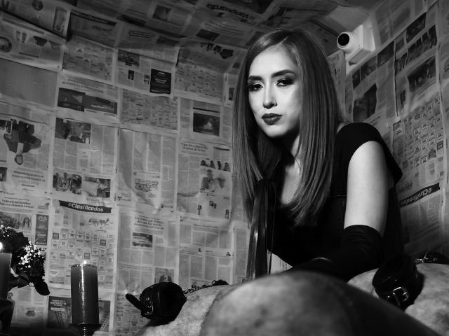 VenusBarret has long, straight, blonde hair, wearing a black top and gloves, with dark lipstick, looking seductively at the camera, surrounded by candles and dark objects in a black-and-white photo.