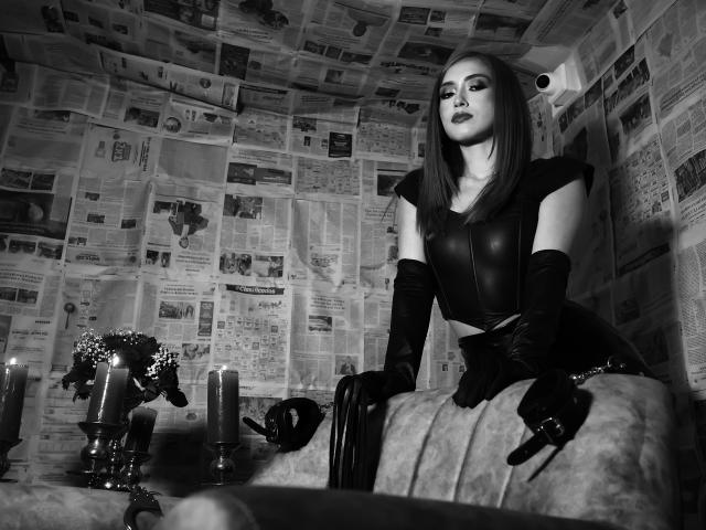 VenusBarret with sleek black leather outfit, long gloves, and dark lipstick, leaning on a couch with candles and flowers. Her tits are small, her ass is hidden. Black-and-white photo.