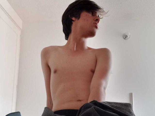 AngelW, shirtless with a slim, pale, and smooth upper body, dark hair, and small nipples, holding a gray towel in a bright room, looking to the side.