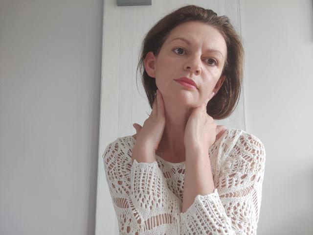 VivianTease has short brown hair, wearing a white crochet top, hands touching her neck, looking slightly to the side with a subtle smile. Her tits are small and visible.