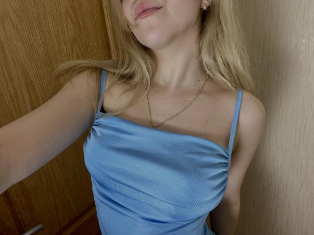 SugarrX, avec ses cheveux blonds, portant une robe en satin bleu, montrant sa poitrine, un collier en or et un maquillage discret, dans une pièce en bois. Ses lèvres sont légèrement entrouvertes..