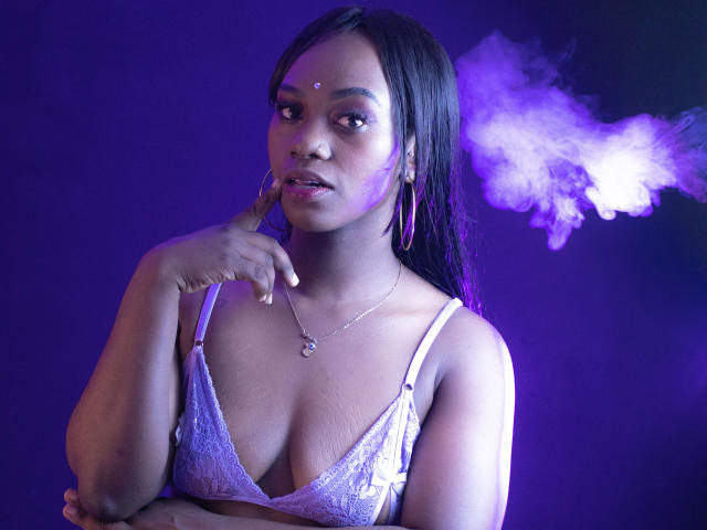 AliceDuperly, à la peau foncée, portant un soutien-gorge en dentelle violet, a un doigt sur ses lèvres, elle fume, avec une lumière violette et bleue, de longs cheveux noirs, des boucles d'oreilles cerceaux et un petit point sur le front..