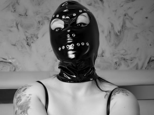 KatalinaJohnson with black latex mask, metal eyelets, and straps, showing tattoos on shoulders, wearing thin-strapped top, black and white photo.