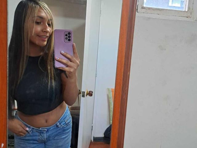 ThaliaMorrison, avec de longs cheveux blonds et noirs, portant un crop top foncé et un jean bleu, tenant un téléphone violet, montrant ses petites seins et ses fesses plates dans un selfie pris devant un miroir..
