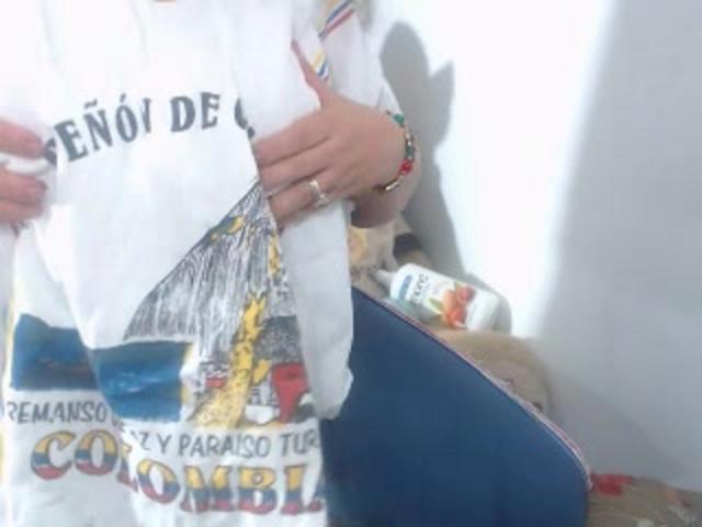 GilliSkquirtty's hands holding a white shirt with colorful print, wearing a red bracelet, blue bag with a yellow pattern, and a ring on the right hand.