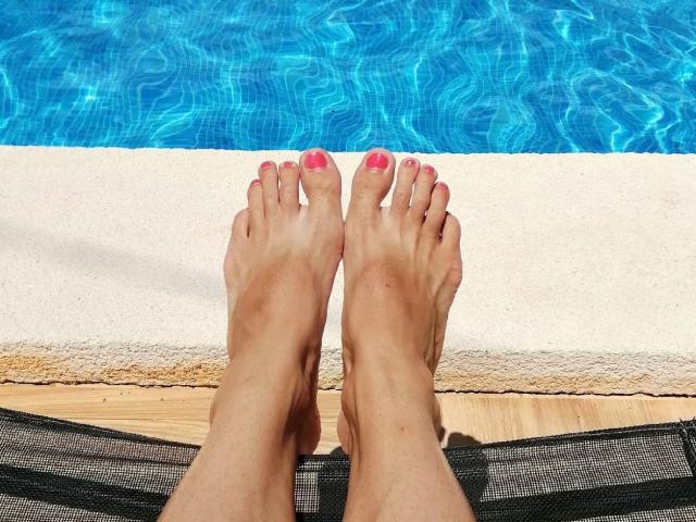 I piedi di LaFrancaise, con le unghie dei piedi dipinte di rosa, le dita leggermente incurvate, poggiati su una poltrona in rete nera, accanto a una piscina luminosa di colore blu, con l'acqua che crea delle increspature..
