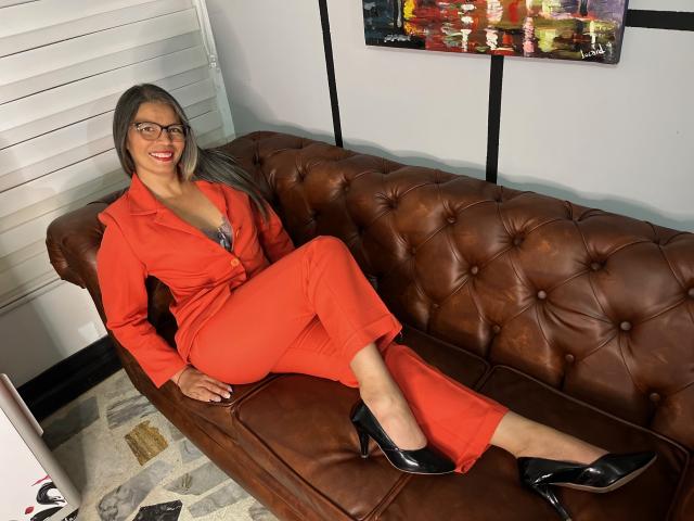 ParisMatur69, con cabello gris, gafas, traje rojo, mostrando los senos, sentada en un sofá de cuero marrón, con tacones negros, sonriendo, con las piernas cruzadas..