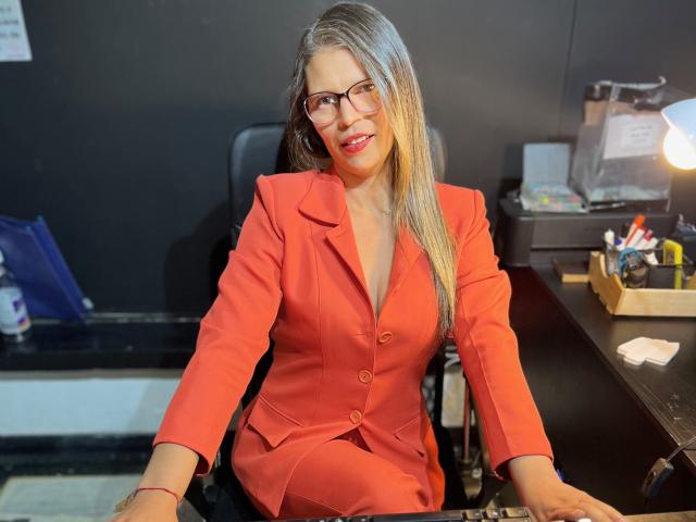ParisMatur69, con lunghi capelli biondi, occhiali e un blazer rosso, seduta alla scrivania, sorridente, con le mani appoggiate sulla sedia nera, parete nera, scrivania con documenti e lampada sullo sfondo..