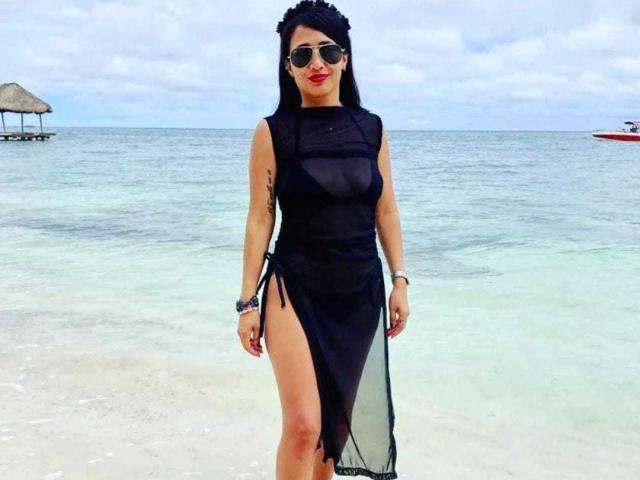 Evangeline in einem schwarzen, transparenten Kleid mit hohem Beinschlitz, der ihre Vulva zeigt, mit einem schwarzen Blumenhaarreif, Sonnenbrille, rotem Lippenstift und Tattoos am Arm, stehend am Strand, mit Wasser im Hintergrund..