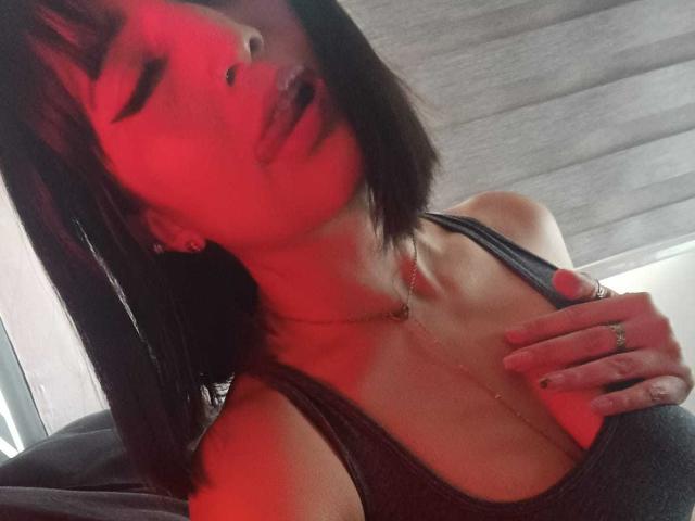 Evangeline met kort zwart haar, een rood aangelopen gezicht, tong uit, een zwarte tanktop, een ketting, hand op de borst, waarbij kleine borsten zichtbaar zijn, een ring aan de vinger, oorbellen, donkere kamer..