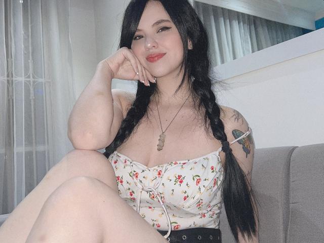 ElyMoon, com longas tranças pretas, vestindo um vestido com alças florais, mostrando os seios, sentada com uma perna levantada, mão no rosto, sorrindo, tatuagem no braço, colar, pele clara..