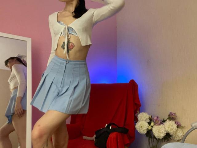 Tsukerberg with a white crop top, blue skirt, and visible tattoo under the tits, standing in front of a mirror, showing a reflection of her ass.