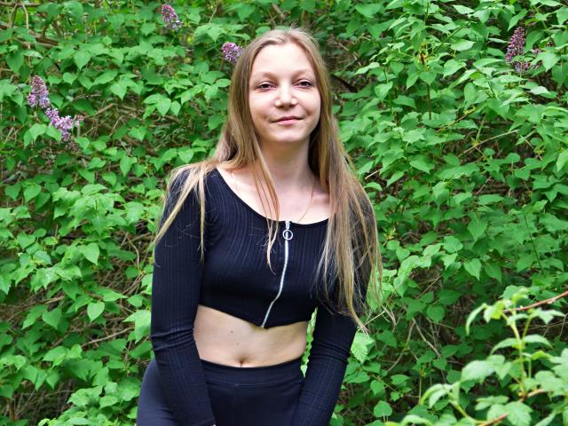 LovelyxEmilly with long blonde hair, wearing a black ribbed crop top and black pants, standing in front of green leafy plants with purple flowers, smiling slightly.
