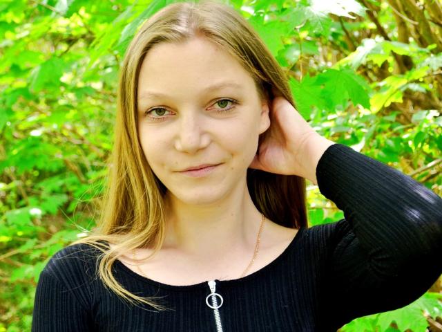 LovelyxEmilly, avec ses cheveux bruns clairs, ses yeux verts et un léger sourire, portant un haut noir texturé avec une fermeture éclair, une main dans ses cheveux, debout dans un feuillage vert..