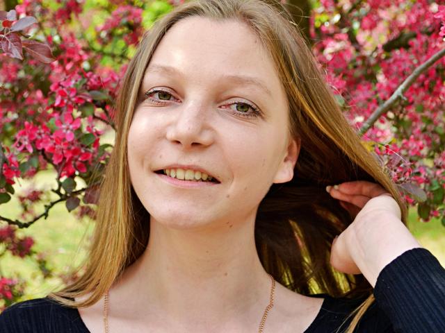 LovelyxEmilly has light brown hair, fair skin, small nose, smiling with visible teeth, wearing a black top, holding her hair, and a gold necklace. Pink flowers in background.