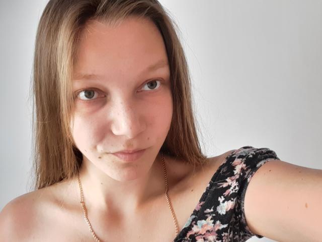 LovelyxEmilly, com cabelos castanhos longos, pele clara, sardas e um nariz pequeno, vestindo uma blusa floral com um ombro de fora e um colar de ouro, sorrindo levemente..