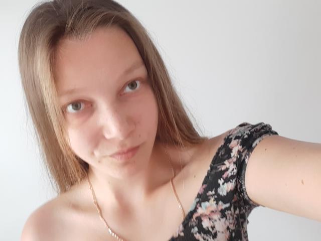 LovelyxEmilly, avec ses cheveux bruns clairs, ses yeux verts et son haut à fleurs avec une épaule dénudée, montrant un petit bout de peau non rasée sur son menton, dans une lumière douce et naturelle..