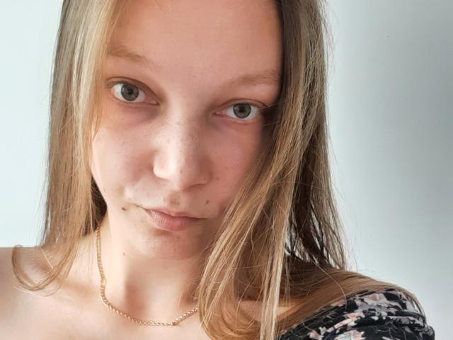 LovelyxEmilly has light brown hair, green eyes, and freckles, with a subtle pout, wearing a floral off-shoulder top and a gold necklace. Her ass is not visible.
