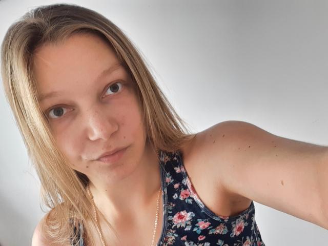 LovelyxEmilly, com cabelo castanho claro, sardas e pequenas manchas no rosto, vestindo uma regata florida, segurando um selfie com uma expressão neutra..