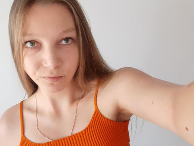 LovelyxEmilly, avec ses cheveux bruns clairs, ses yeux verts, et son débardeur orange côtelé, montrant ses petites seins fermes, et un collier en argent, se prend en selfie avec une légère moue..