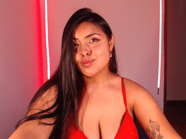 Angela Roggers, avec ses longs cheveux noirs, son soutien-gorge rouge et un tatouage sur son bras droit, sourit légèrement, avec une lumière rouge derrière elle. Ses seins sont de taille moyenne et elle est jolie..