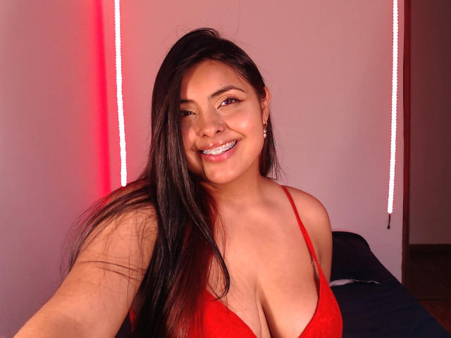 AngelaRoggers met lang zwart haar, lachend, draagt een rode bh, toont haar grote borsten, rode lichtstrips op de achtergrond, maakt een selfie..