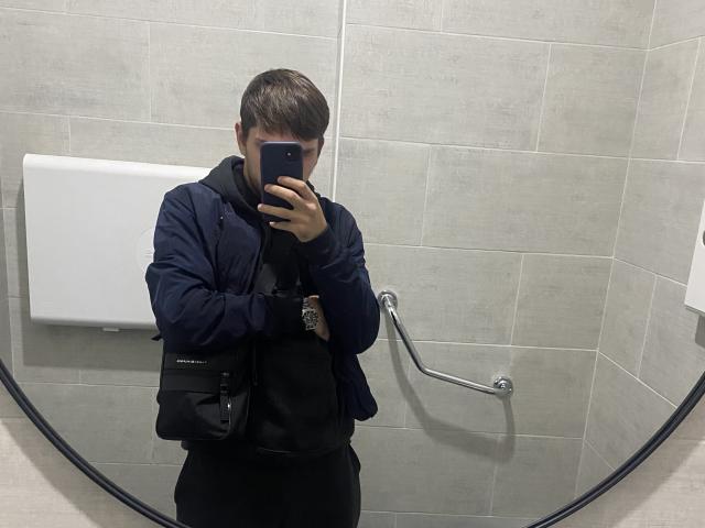 SashaaLovee, con i capelli castani, indossa una giacca blu scuro, una maglietta nera e pantaloni neri, e scatta un selfie allo specchio in un bagno, tenendo in mano un telefono e mostrando una mano..