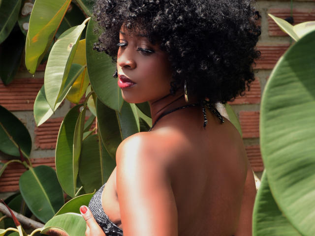 KarmenCastillo with curly black afro, dark skin, red lipstick, wearing a black halter top, showing her back and small, bare ass, surrounded by green leaves.