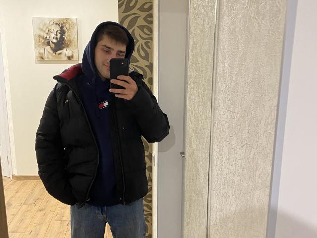 SashaaLovee indossa una giacca nera imbottita, una felpa blu e dei jeans, e sta scattando un selfie allo specchio con un telefono nero, mostrando un'espressione leggermente infastidita, in una stanza con pavimento in legno..