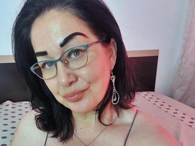 LexyRose has black hair, blue glasses, and a slight smile with her tits partially visible, wearing a chain earring, and has a shiny face with pink light.