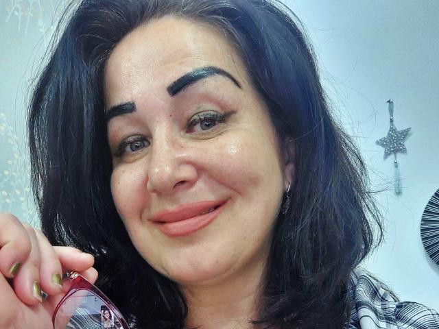 LexyRose with dark, wavy hair, thick eyebrows, and pink lips, holding a can, smiling with a slightly raised ass. She has green nail polish and a star-shaped decoration.