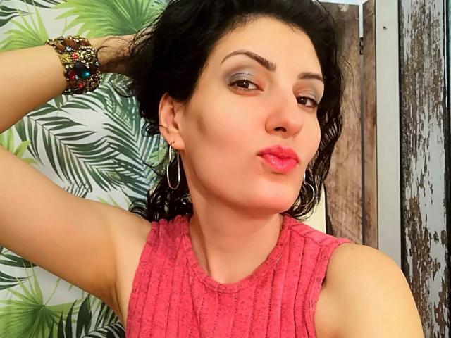AddictedToUTS with pink lips, brown eyes, and wavy black hair, wearing a pink sleeveless top, hoop earrings, and colorful bracelets, puckering for a kiss.