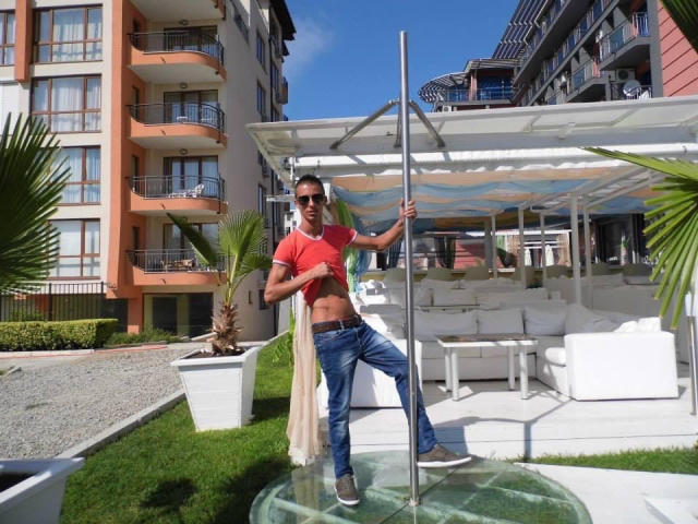 TwinkMario mit schlanker, muskulöser Statur, trägt ein rotes T-Shirt, blaue Jeans und eine Sonnenbrille und steht neben einem Pfosten. Er hebt sein Hemd, um seine trainierten, behaarten Bauchmuskeln zu zeigen..