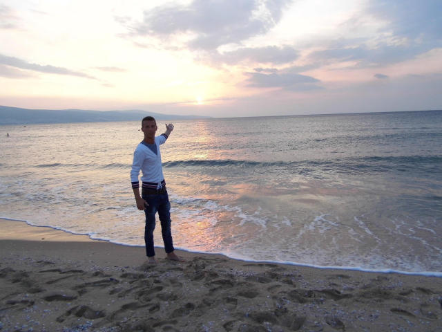 TwinkMario stands on a sandy beach, waving with one hand, wearing a white long-sleeve shirt and blue jeans, at sunset with gentle waves and a cloudy sky.