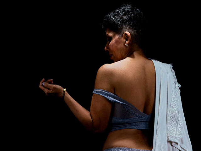 JuliaNewell with short curly black hair, wearing a blue backless top, white sari, and earrings, looking over her shoulder, showing her back and butt, dark background.