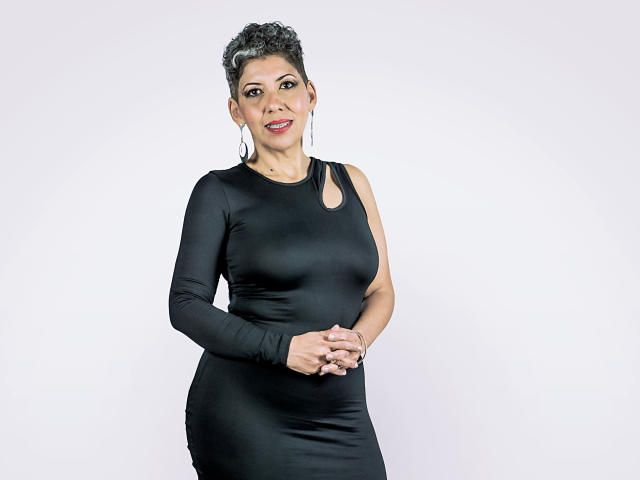 JuliaNewell, short curly black hair, wearing a tight black dress, showing her tits, hands clasped, smiling, hoop earrings, white background.