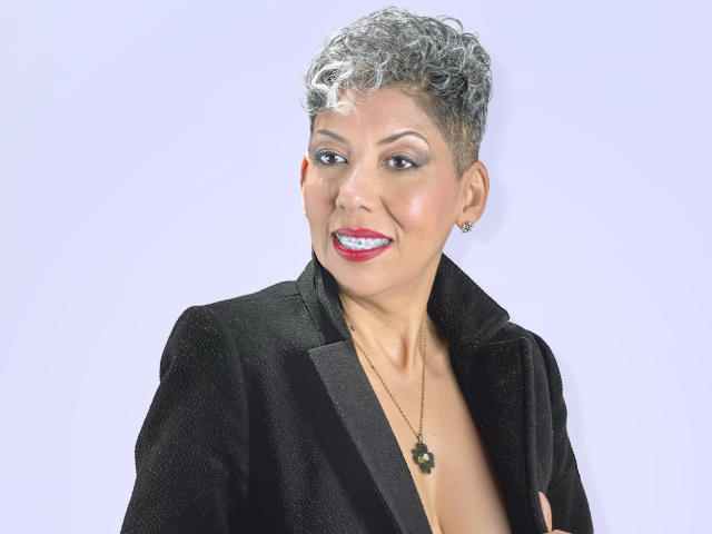 JuliaNewell with short, curly gray hair, black jacket, red lipstick, smiling, wearing a necklace with a black flower, and small earrings, showing her tits under the jacket.