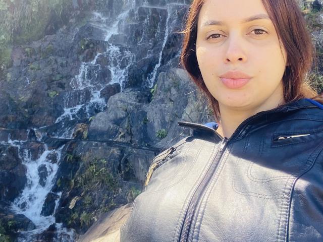 EmilyParkerr with puffy pink lips, brown eyes, and short brown hair, wearing a gray zip-up jacket, standing in front of a rocky waterfall. Boobs visible under jacket.