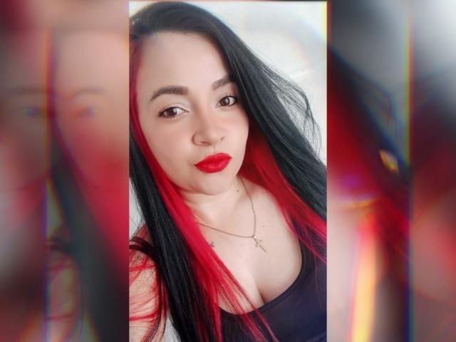 PAMDORA mit langem, schwarzem Haar und roten Strähnen, trägt roten Lippenstift und ein schwarzes Tanktop, wobei ihre Brüste zu sehen sind und sie eine Kreuzkette trägt. Sie lächelt in die Kamera..