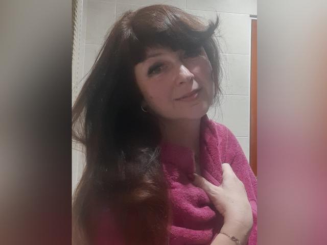 Bianca: Eine Frau mit langen, braunen Haaren, die ein pinkfarbenes Handtuch trägt, ihre Brust berührt, leicht lächelt und in die Kamera schaut. Sie befindet sich in einem Badezimmer und hat einen subtilen, sexy Ausdruck..