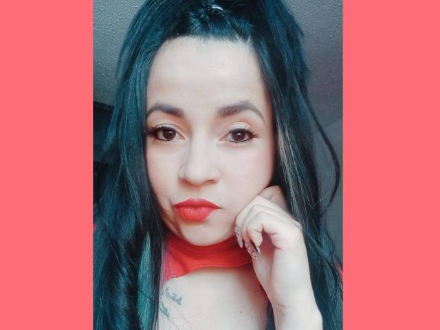 PAMDORA with long black hair, red lipstick, and subtle makeup, pouting with hand on cheek, showing tattoo on upper chest, against a red background.