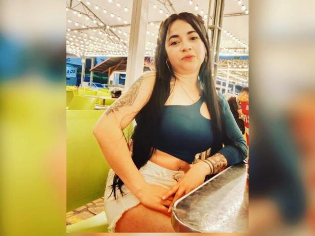PAMDORA mit langen, schwarzen Haaren, einem blauen Crop-Top, weißen Shorts, Tätowierungen am Arm, sitzt an einem Tisch, eine Hand am Oberschenkel, blickt in die Kamera, Innenaufnahme, warmes Licht..