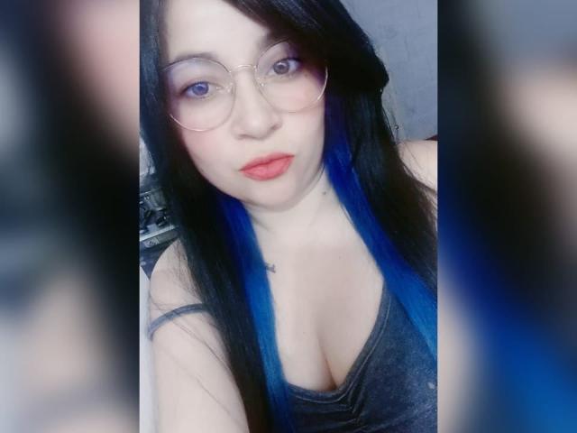 PAMDORA with blue-dyed black hair, glasses, and red lipstick, showing cleavage in a grey top, has pale skin and a cute, sexy face. Boobs are prominent.