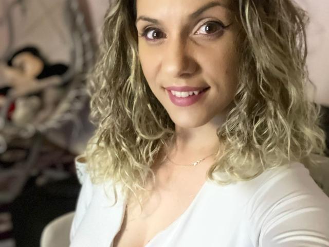 LucilleXSugar mit welligem, blondem Haar, rosa Lippenstift, weißem Oberteil, lächelnd in die Kamera, wobei sie ihre Brüste leicht durch den tiefen Ausschnitt sichtbar macht, trägt eine zarte Halskette..