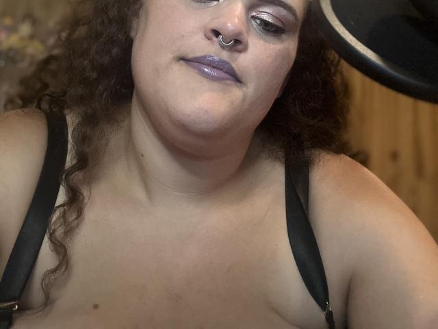 Yvannah met krullend bruin haar, paarse lippenstift, een neusring, zwarte bandjes, die naar beneden kijkt, licht glimlachend, waarbij haar blote borst en kleine borsten zichtbaar zijn..