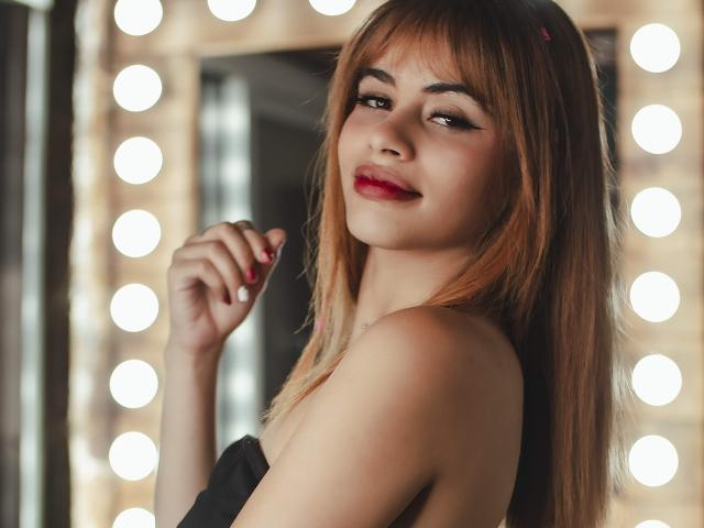 Zenda Evans, met lang bruin haar, rode lippenstift en een zwarte top zonder bandjes, glimlacht subtiel, met haar hand in de buurt van haar kin. Zacht licht van ronde spiegellampen, die haar billen en borsten laten zien..