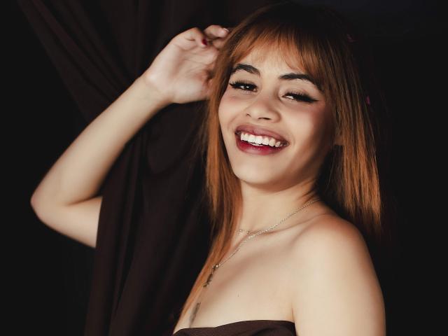 ZendaEvans with brown hair, smiling, showing white teeth, wearing a strapless brown top, touching her hair, wearing a necklace, with visible cleavage and light skin.